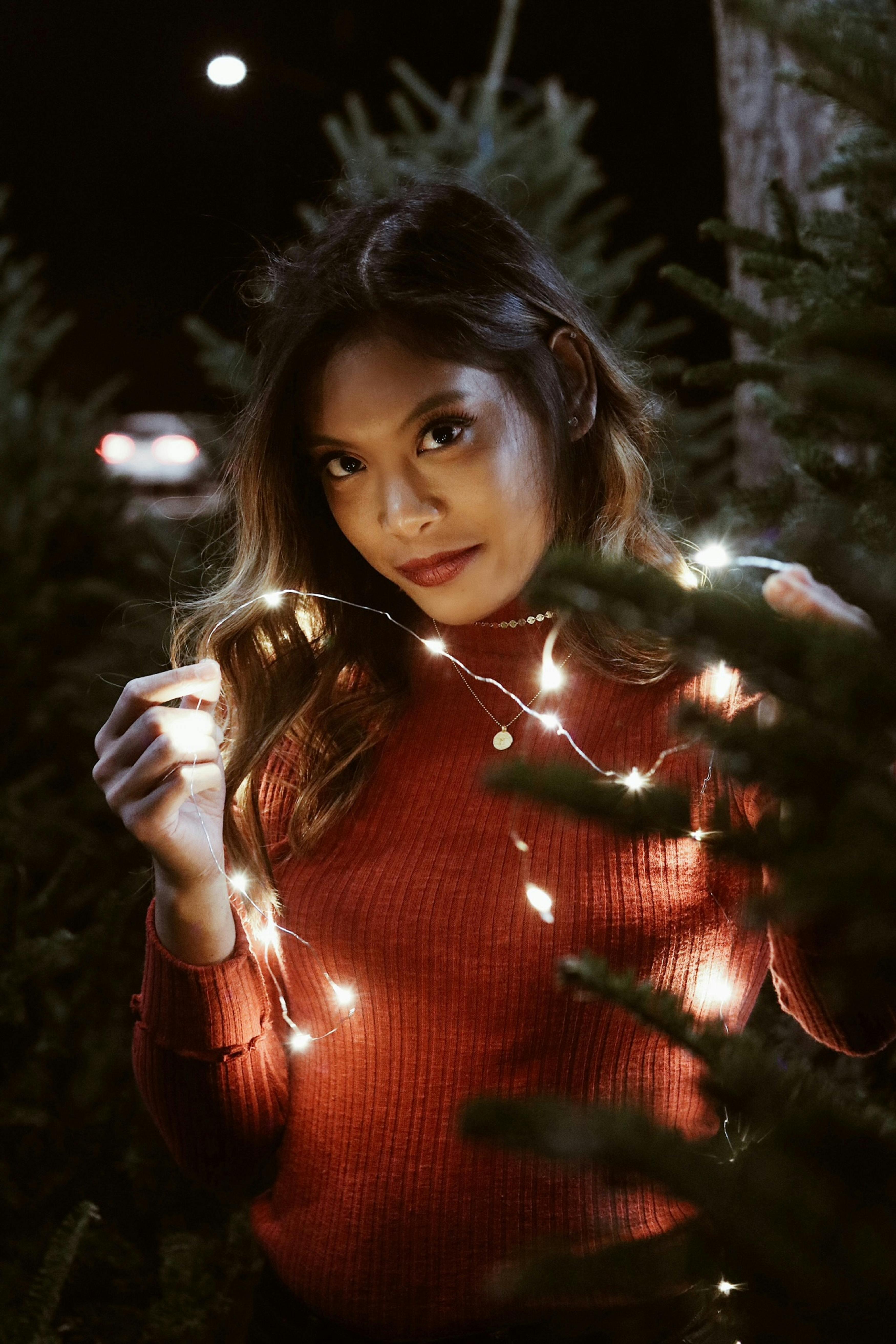 Young woman posing with festive string lights surrounded by Christmas trees at night.