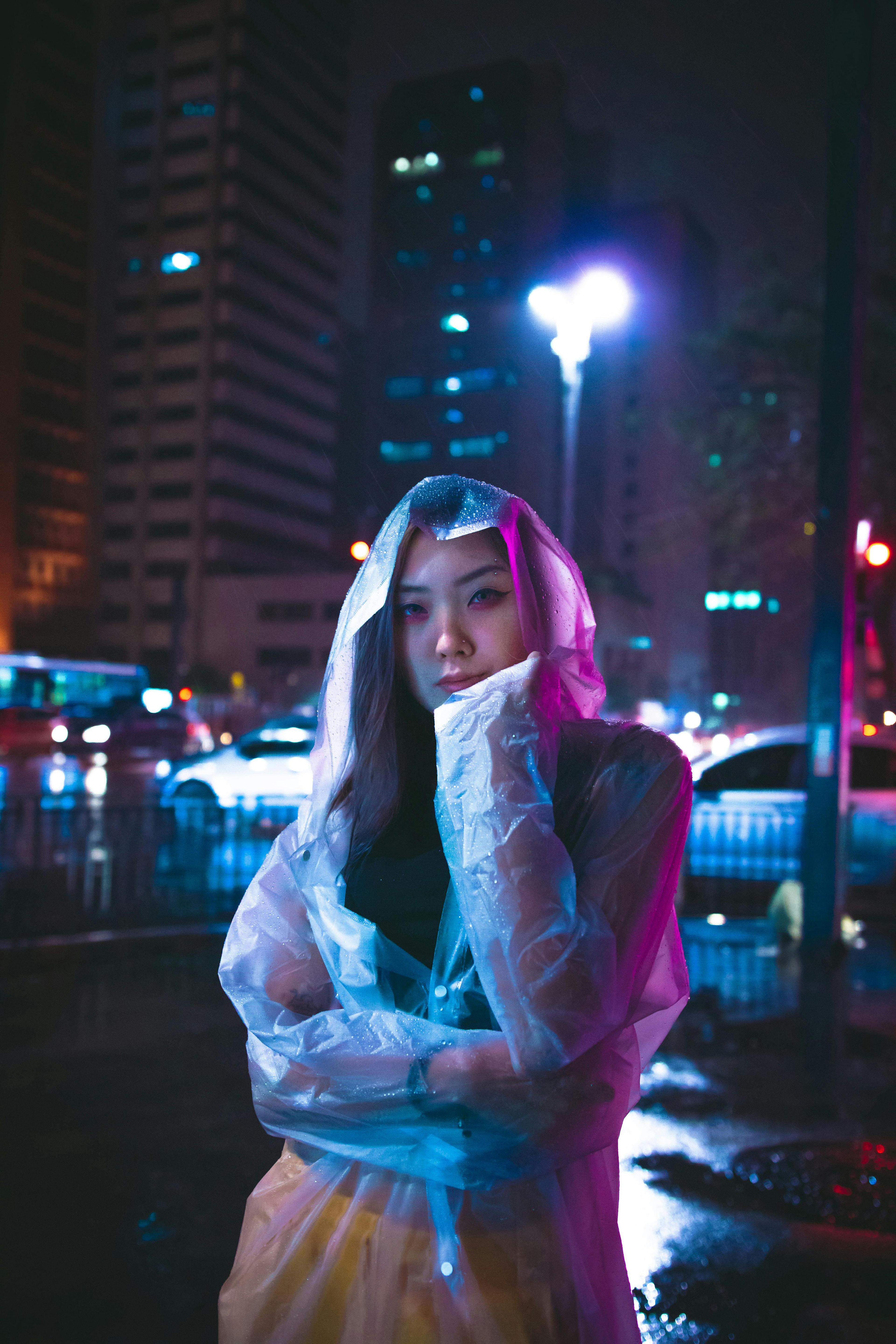 Woman in raincoat poses on city street at night with colorful lights and reflections.