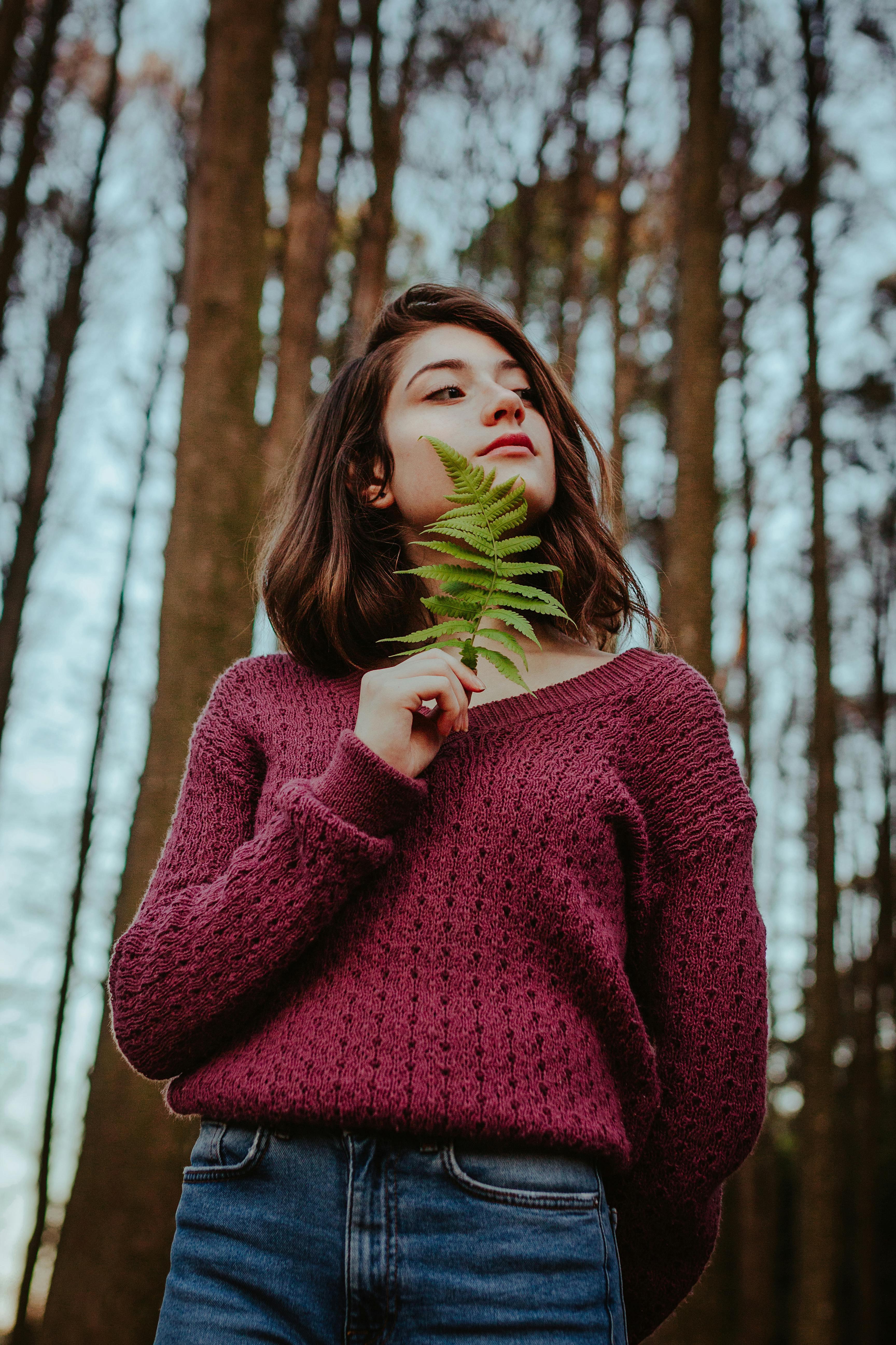 Portrait of a woman in a sweater holding a fern leaf in a serene forest setting.