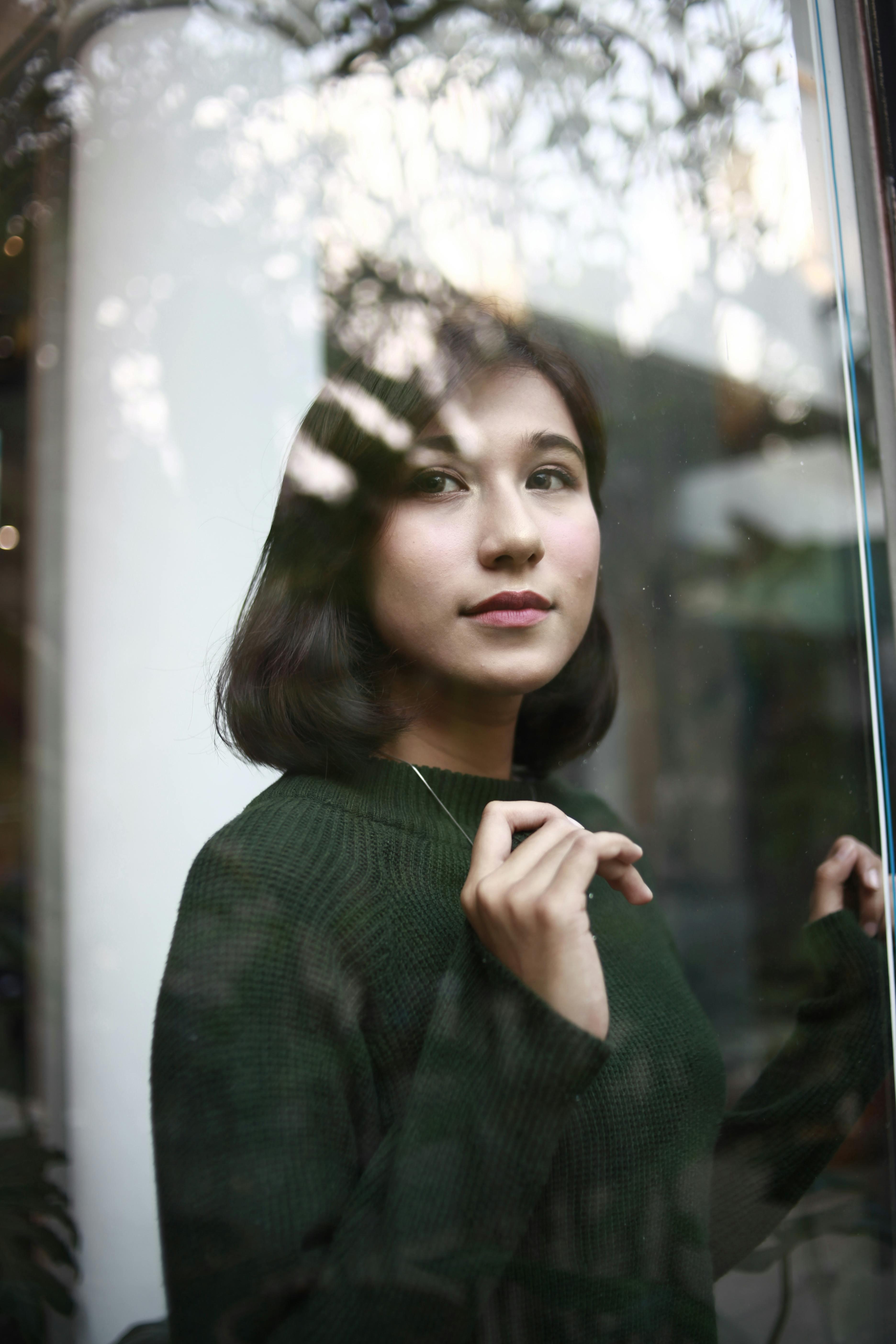 Close-up portrait of a woman in a green sweater gazing thoughtfully through a window.