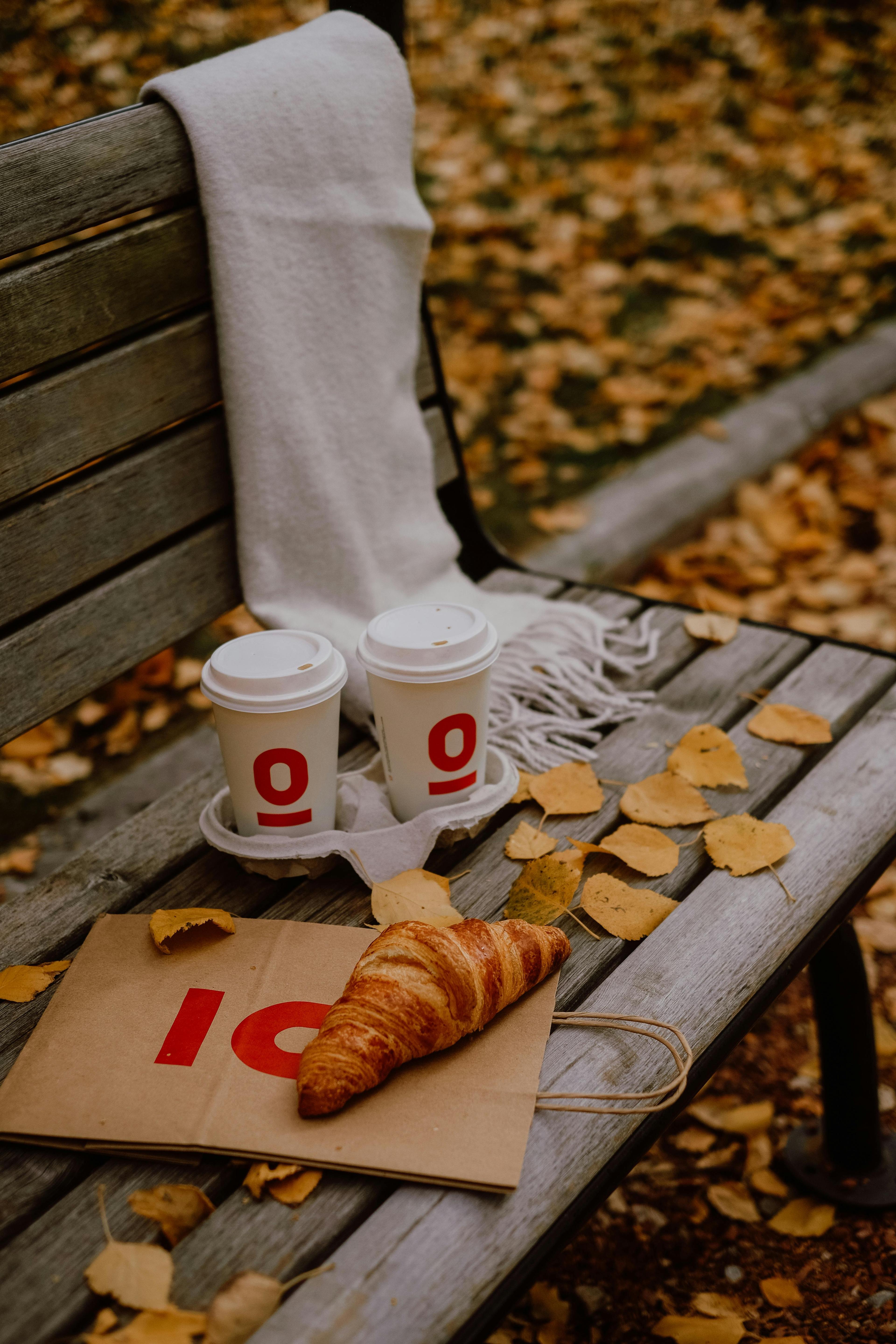 Cup of coffee and a croissant on a bench amid autumn leaves, exuding warmth and relaxation.
