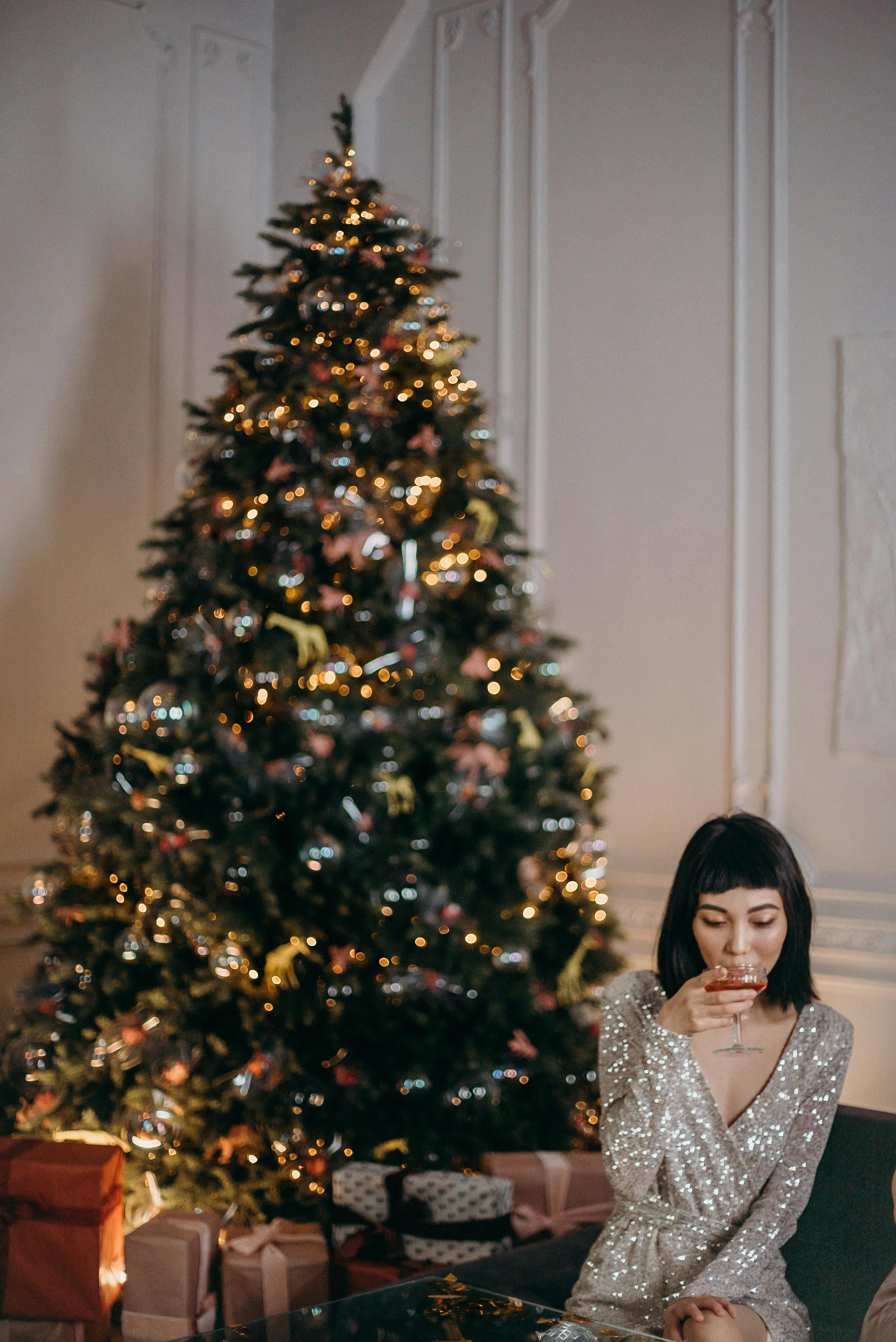 Woman in silver dress enjoying a festive drink by a decorated Christmas tree indoors.
