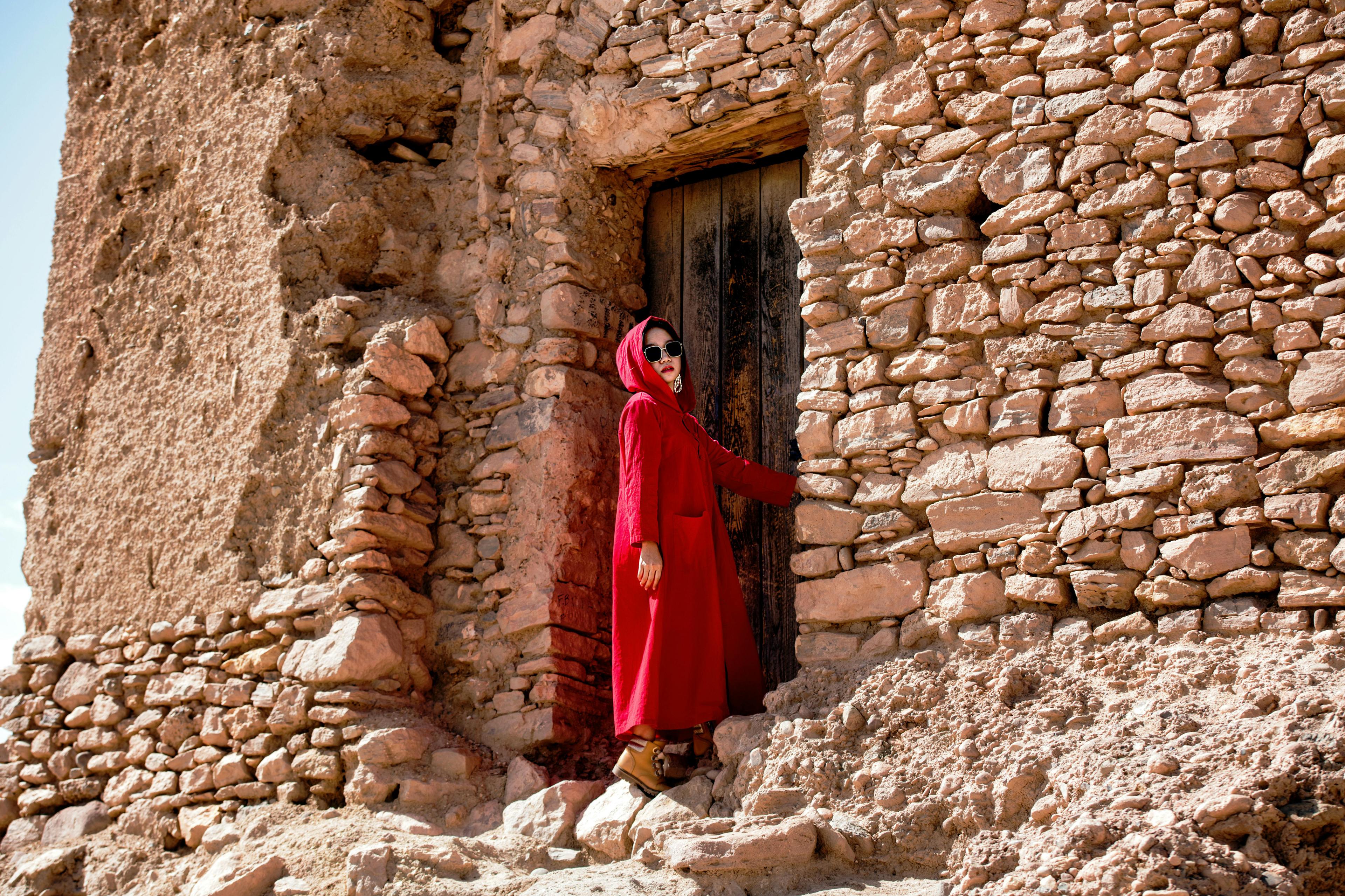 Woman in a red coat standing by a rustic stone wall under bright sunlight.