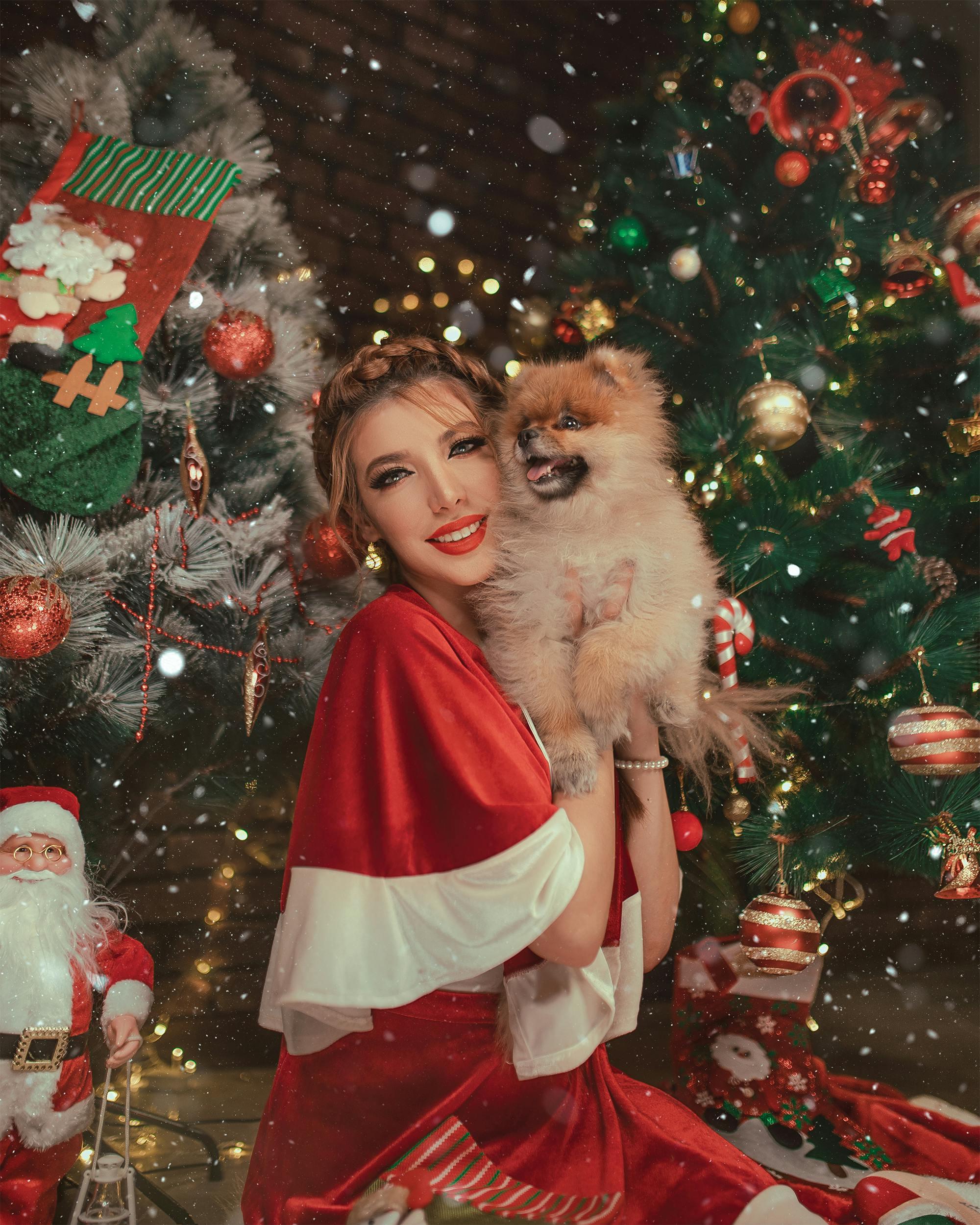 Woman in Santa outfit smiling with a dog in front of decorated Christmas tree.