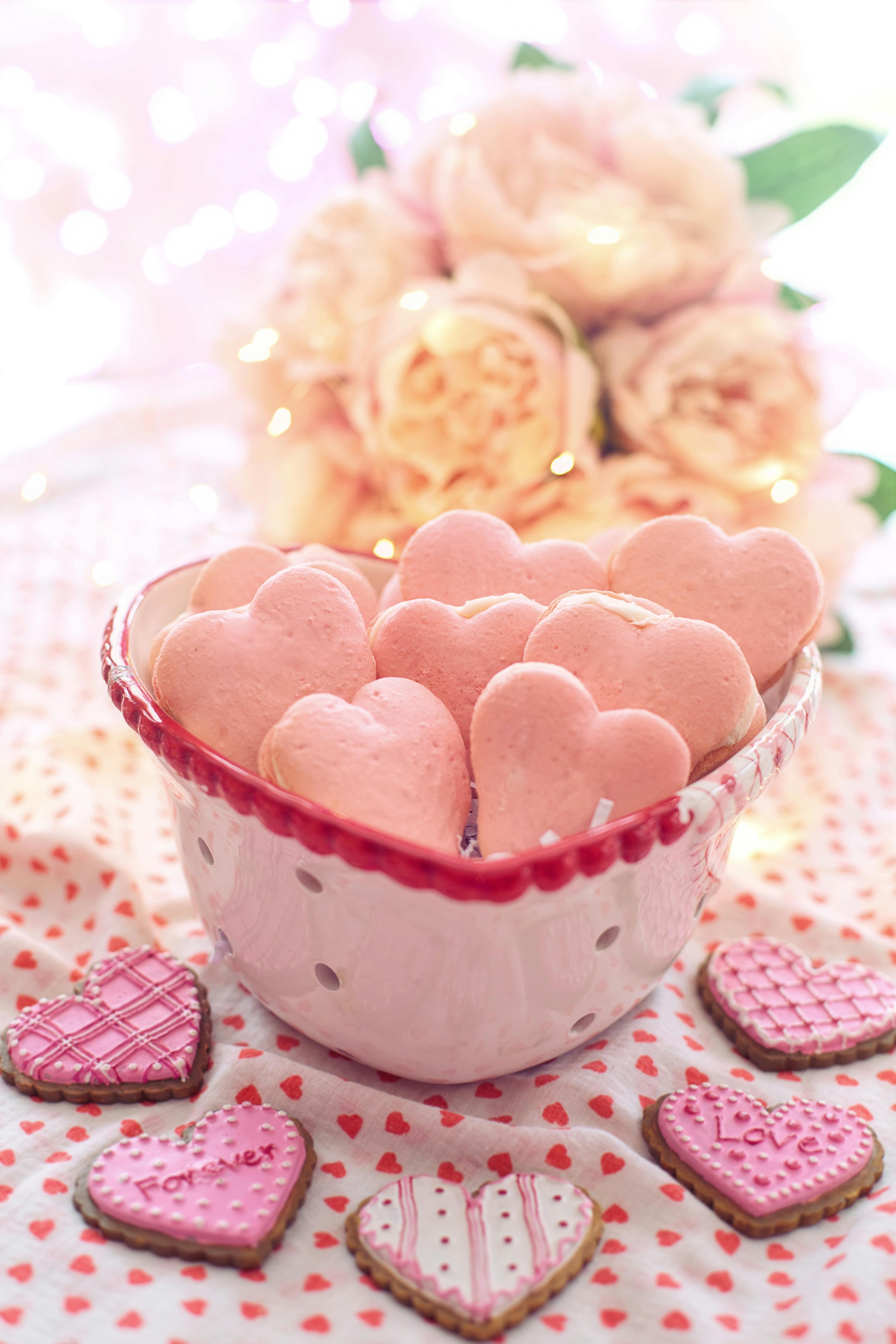 Heart-shaped pink macarons in a bowl with roses, perfect for Valentine's Day treats.