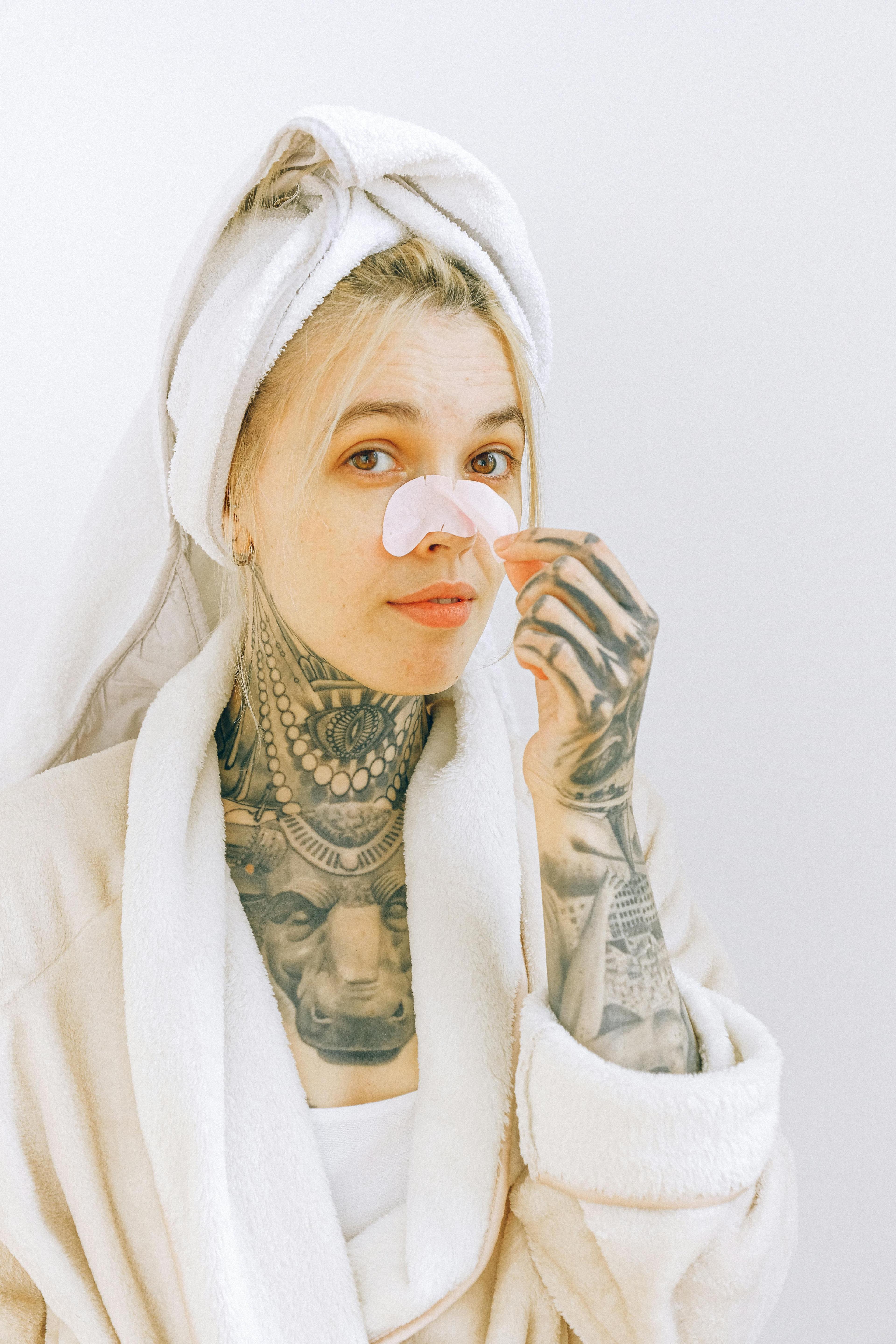 Young tattooed female in terry robe and towel taking off cosmetic nose mask while standing near white wall and looking at camera