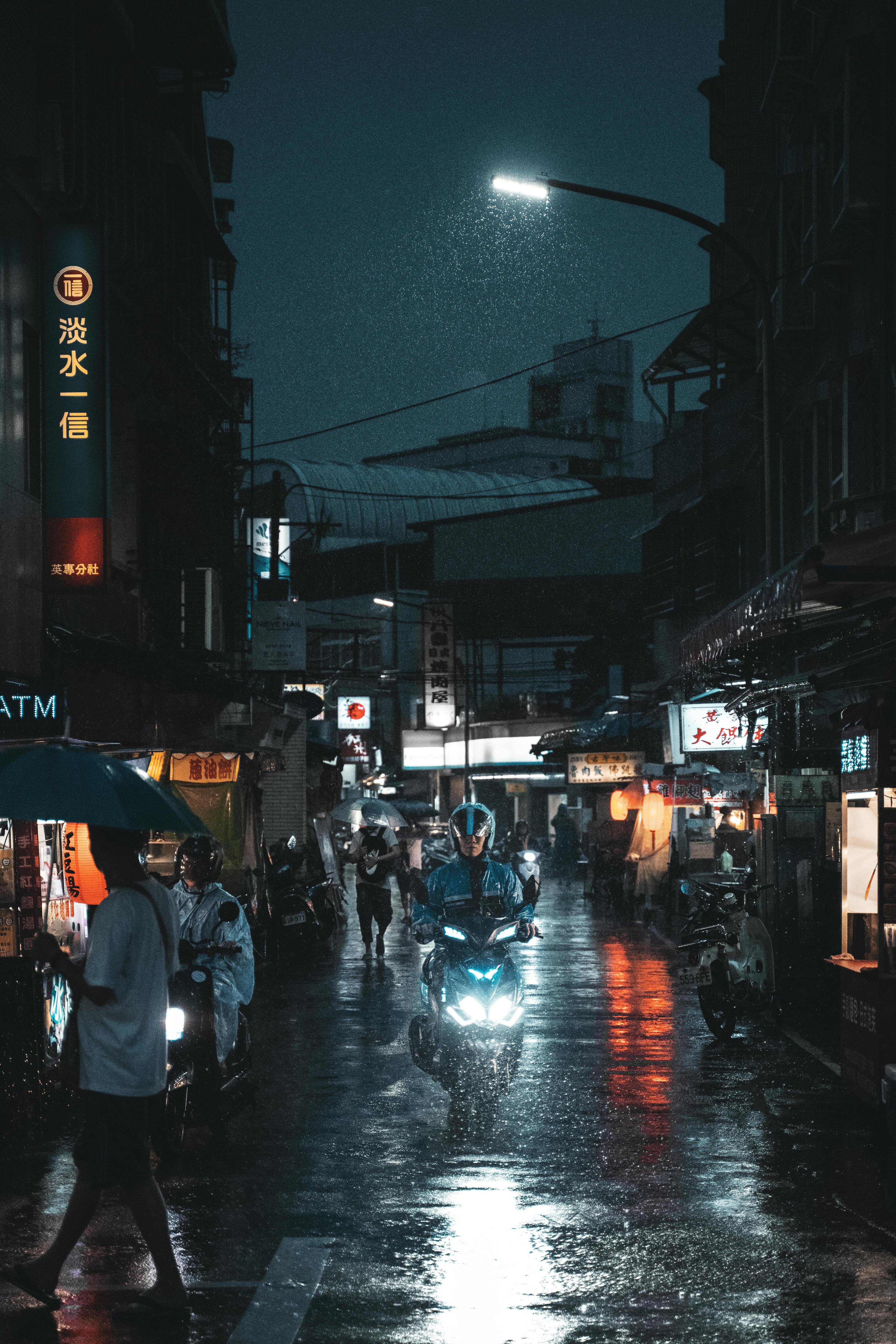 Anonymous motorcyclist in helmet riding bike with headlight on while passing on asphalt roadway illuminating by artificial light of lamp post under people walking on crosswalk in town in twilight