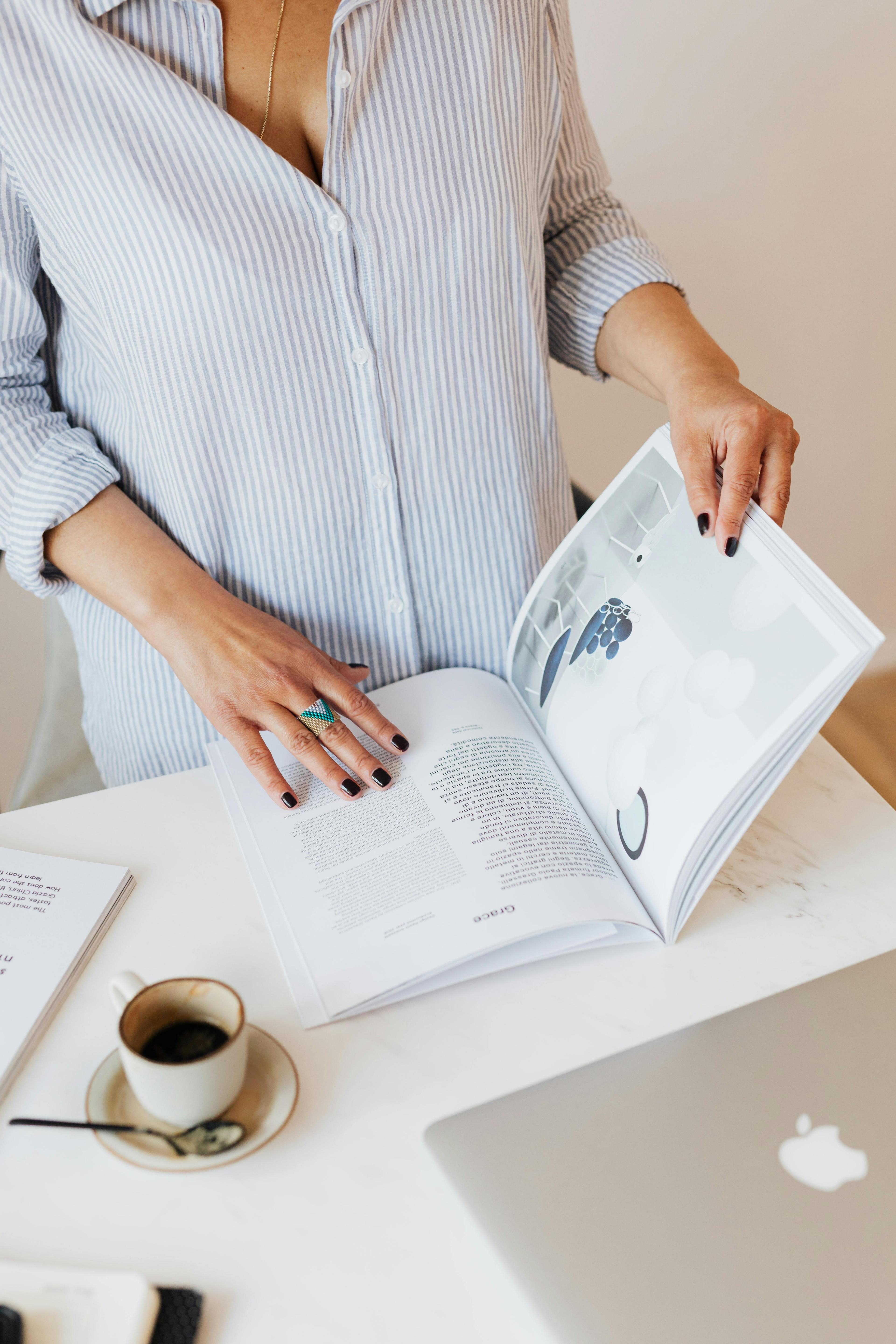 From above crop anonymous female in blue shirt standing near desk with laptop and cup of fresh coffee while turning over professional journal against white wall