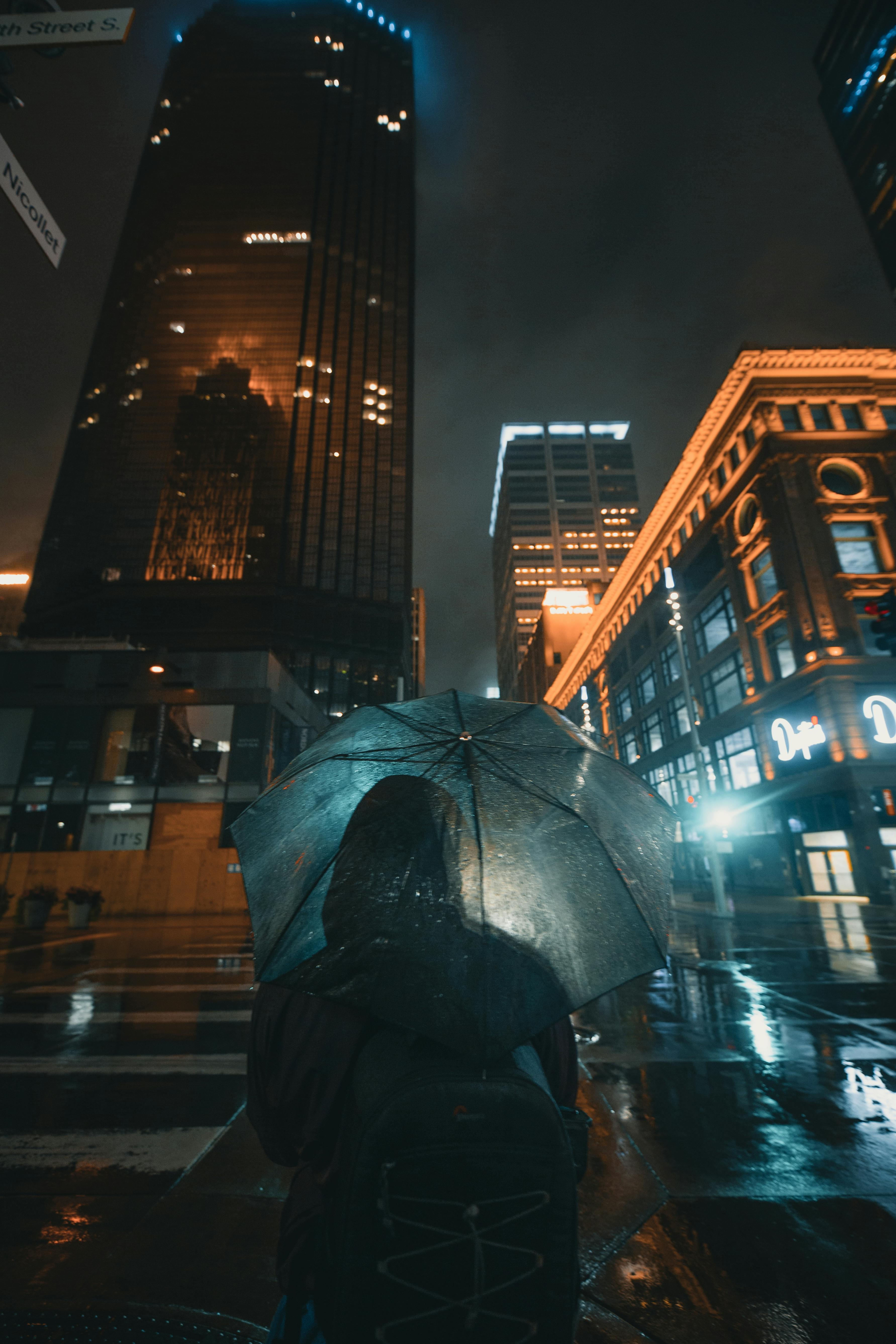 A person in silhouette with an umbrella under illuminated skyscrapers in Minneapolis's wet streets at night.