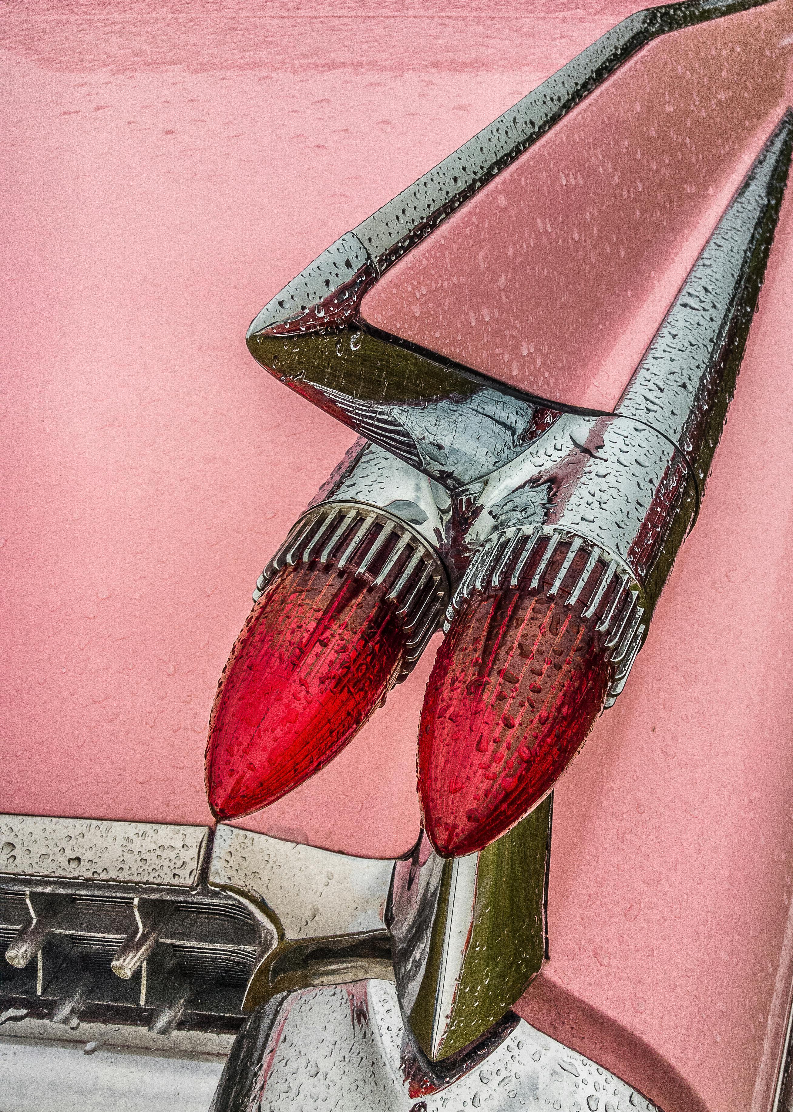 A detailed view of a vintage pink car's chrome tail fins and red lights, covered in water droplets.