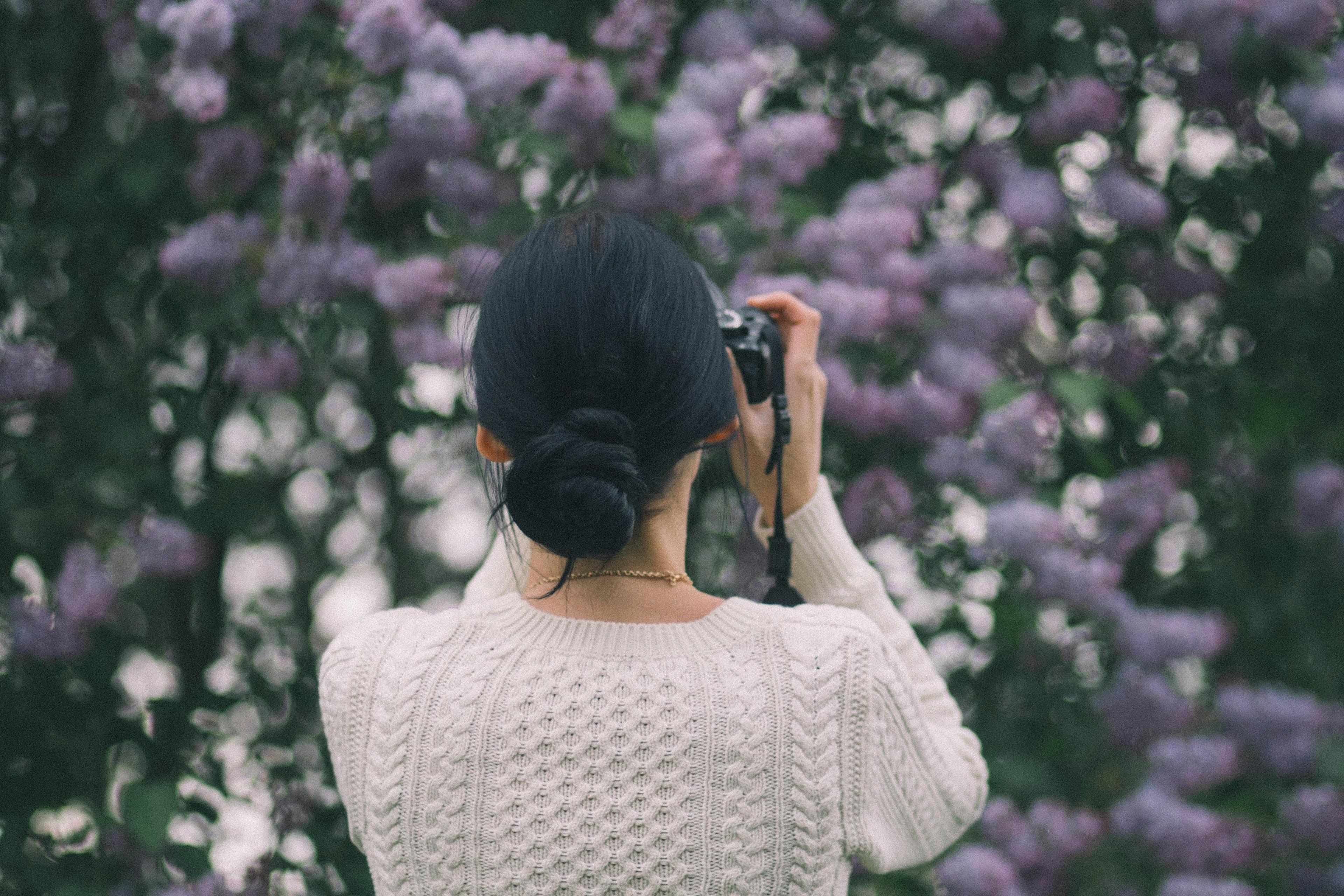 A woman with braided hair capturing vibrant lilac flowers in an outdoor garden during the day.